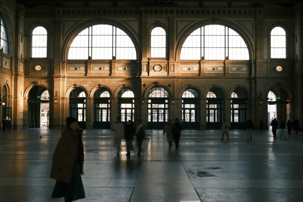 People walking through a grand, sunlit train station hall.